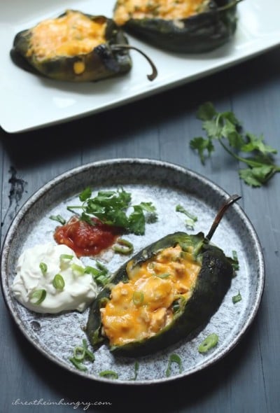 Keto Cheesy Chicken Stuffed Poblanos on a plate with sour cream and salsa garnish, with a platter of poblanos in the background