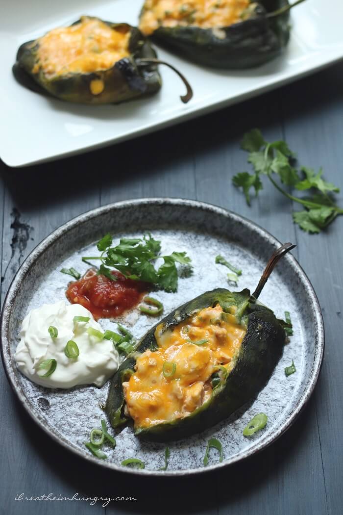 Keto Cheesy Chicken Stuffed Poblanos on a plate with sour cream and salsa garnish, with a platter of poblanos in the background