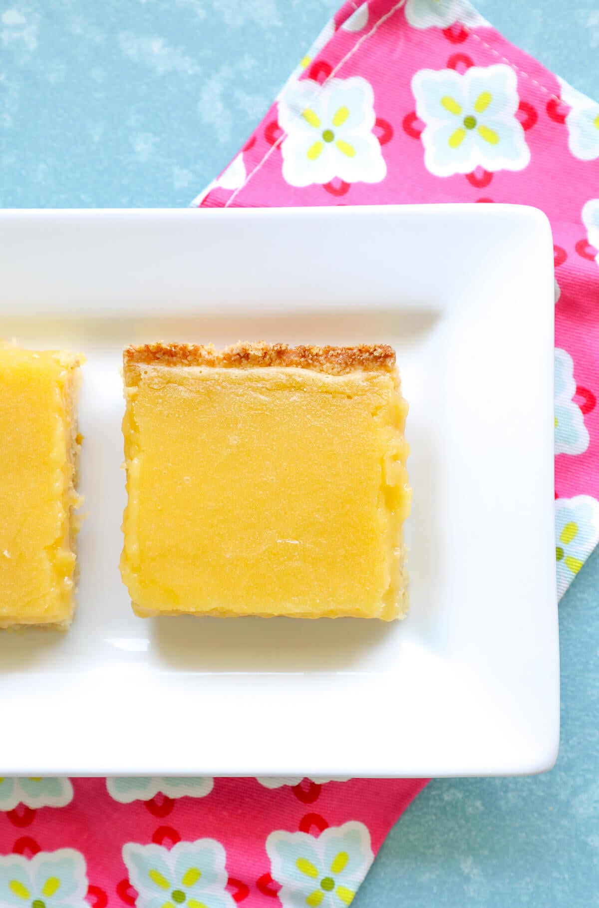 Top view of a lemon square on a white plate with a pink and blue napkin underneath it.