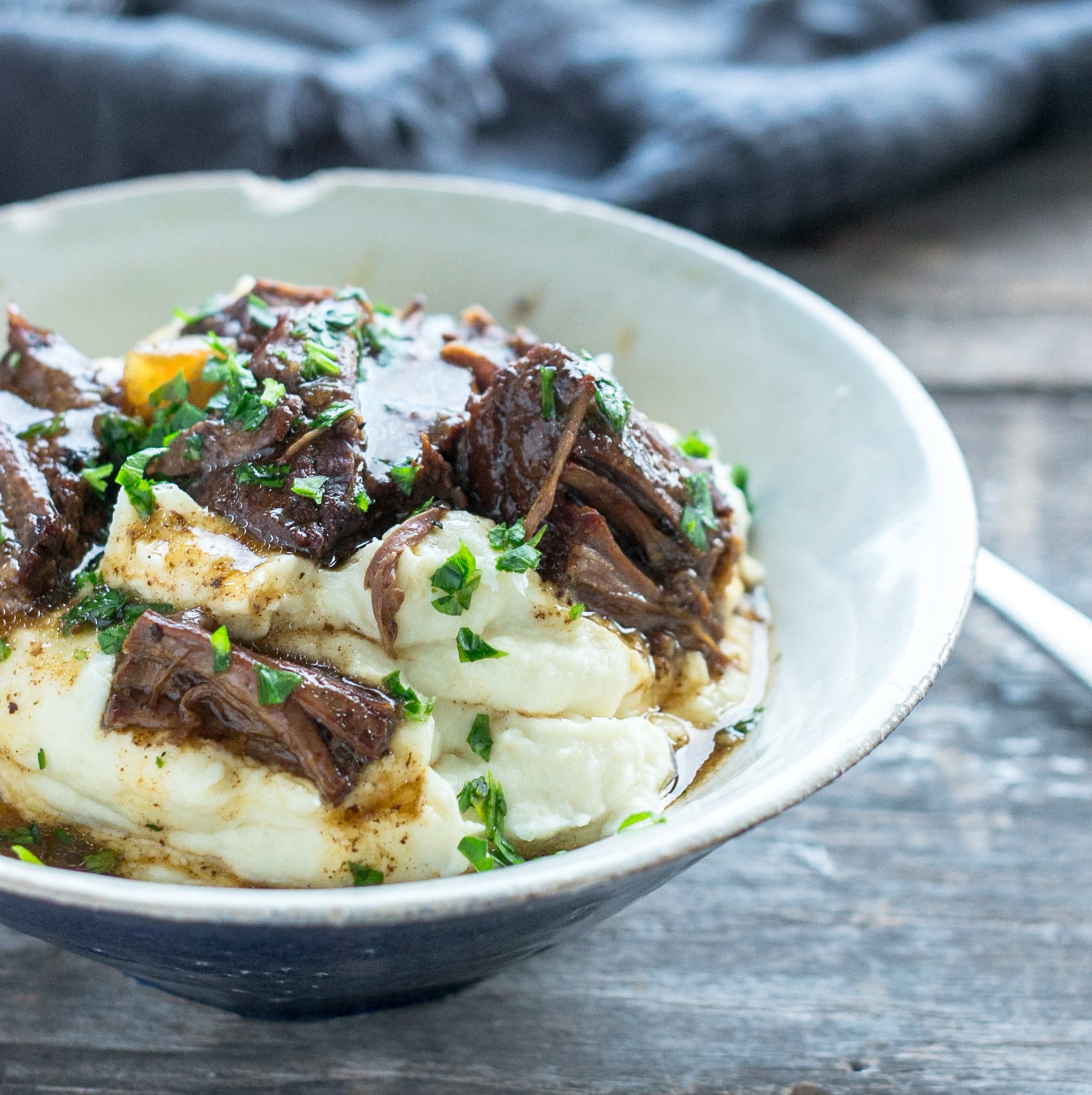 side view of chunks of pot roast in a blue and white bowl with cauliflower puree and garnished with chopped parsley