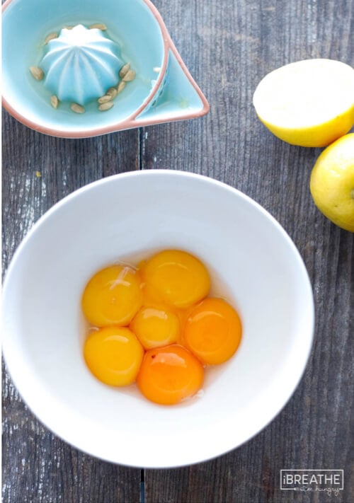 6 egg yolks in a white bowl with lemons and a blue pottery citrus juicer on a wooden board in the background