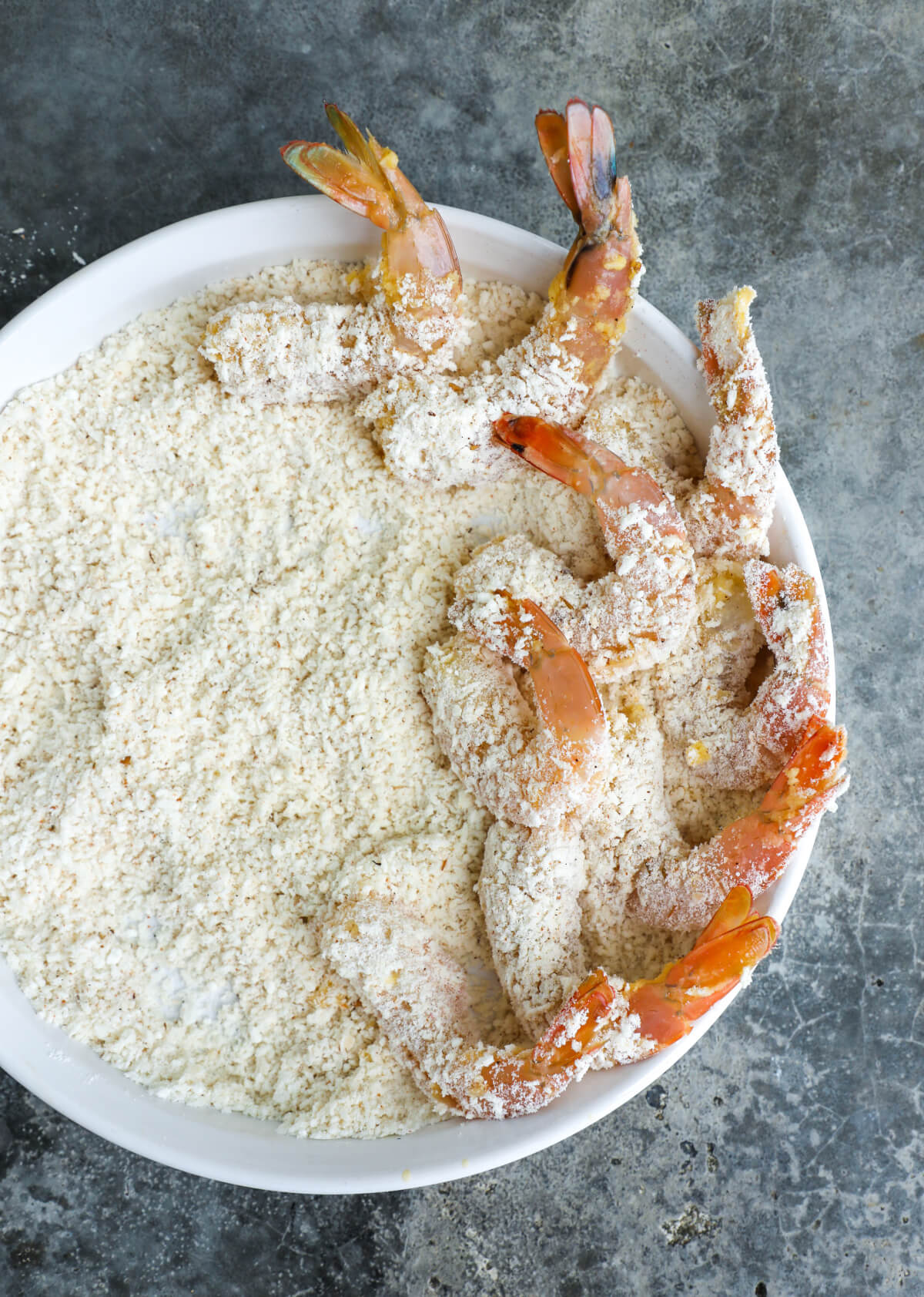 Raw shrimp with coating on it sitting on a bed of breading in a white bowl arranged on the right side.