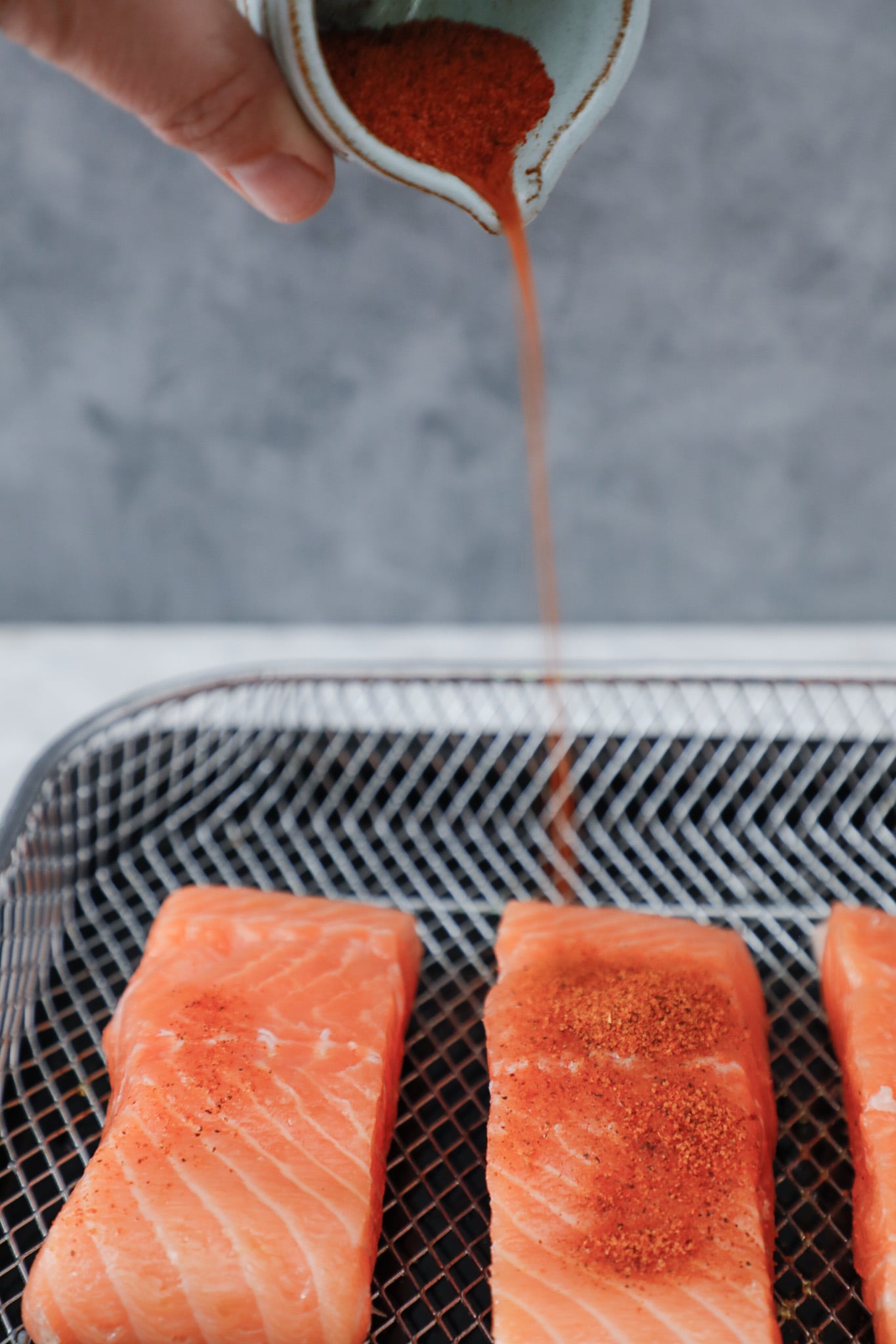 salmon fillets in an air fryer basket with cajun seasoning being sprinkled over it before cooking.