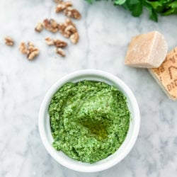 garlic scape pesto in a white bowl on a marble table. Surrounded by parmesan, walnuts and parsley arranged around the bowl