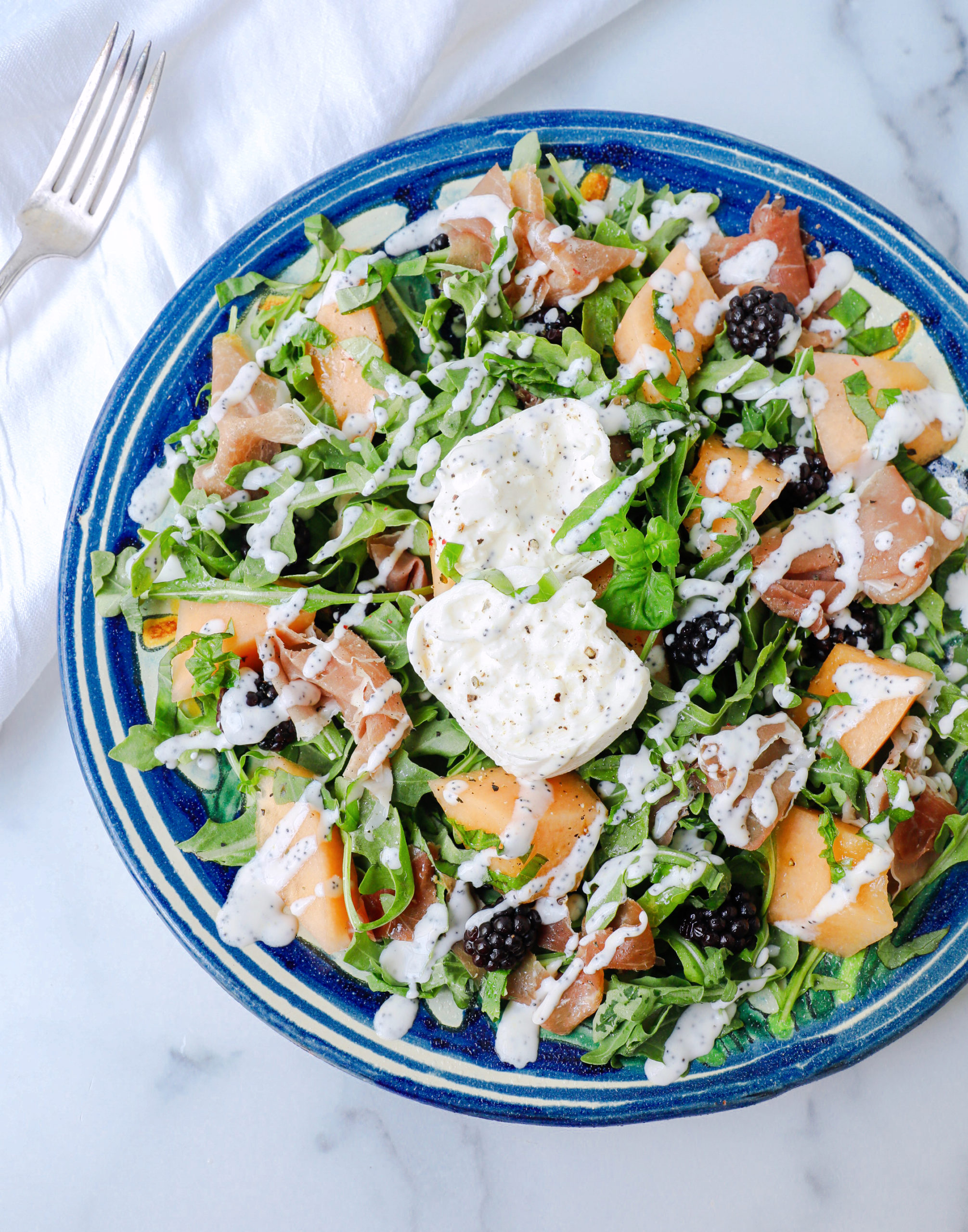 arugula salad with cantaloupe, blackberries, burrata and proscuitto on a blue pottery platter drizzled with lemon poppyseed dressing.