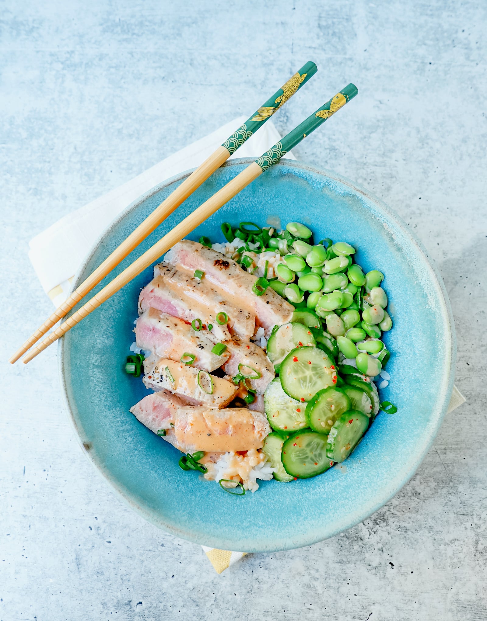 tuna, rice, cucumbers, and edamame in an aqua pottery bowl with chopsticks laying across the top on a diagonal.