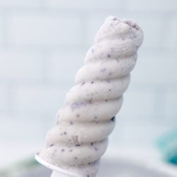 A single blueberry cheesecake protein popsicle held in the air with a white subway tile background
