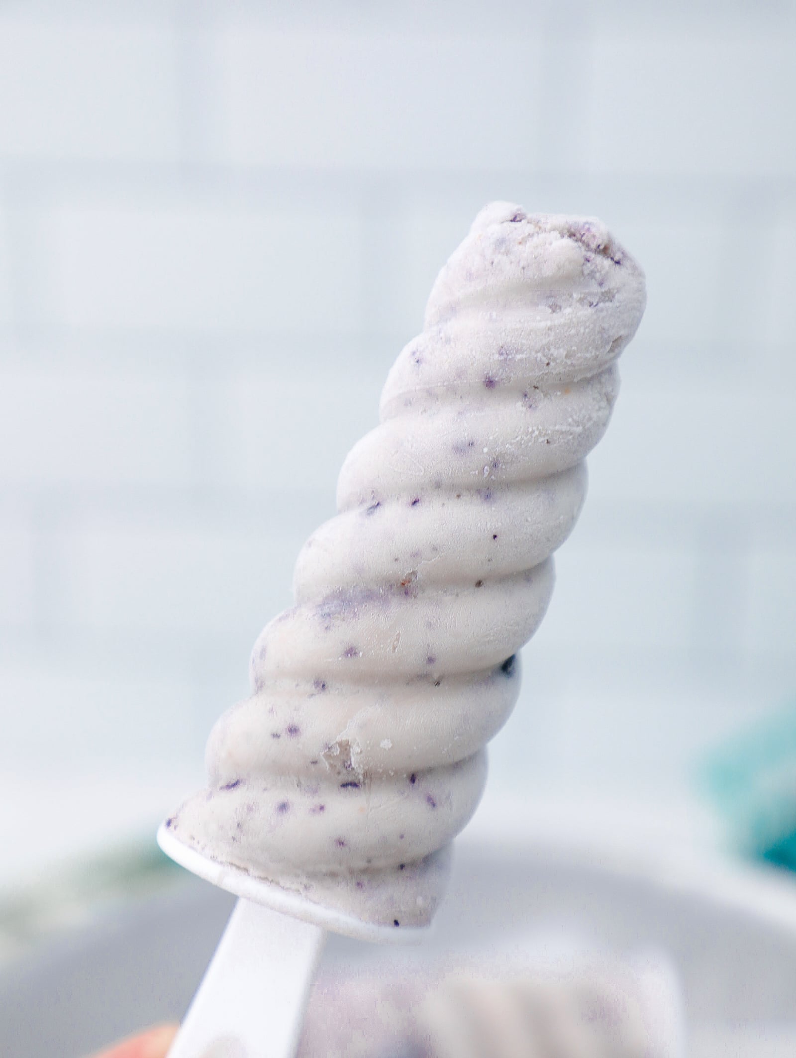 A single blueberry cheesecake protein popsicle held in the air with a white subway tile background