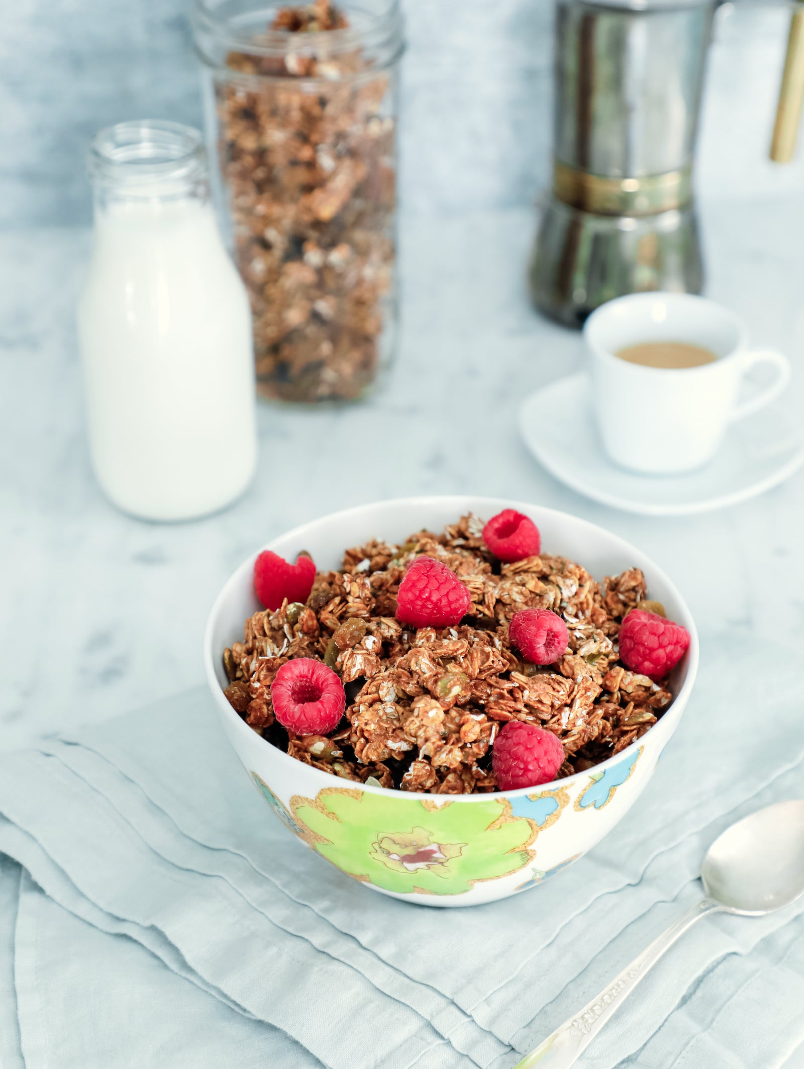high protein granola in a bowl with fresh raspberries on top. in the background is an espresso, bottle of milk, and a canister of the same granola.