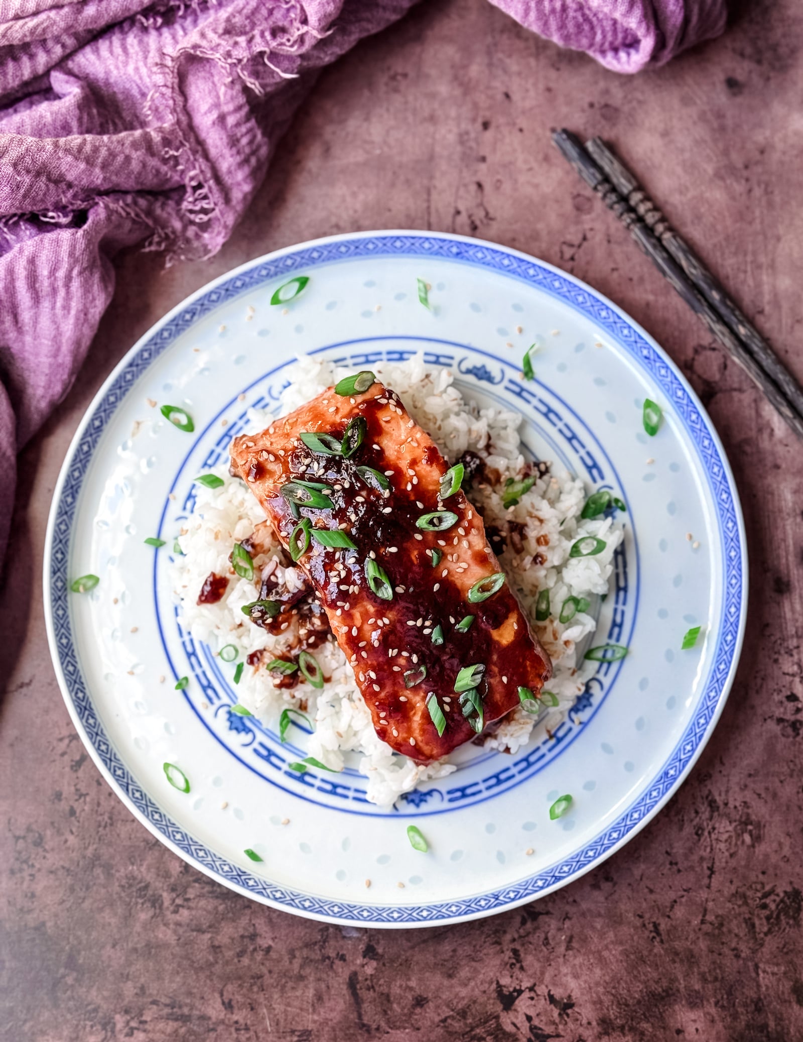 Top view of a salmon filet on a bed of sticky rice, drizzled with hoisin butter and garnished with sesame seeds and sliced scallions. Served on a white rice china plate with blue markings. wooden chopsticks on the top right corner of the plate.