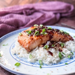 Hoisin Butter Salmon on a bed of white rice, garnished with toasted sesame seeds and sliced scallions. In the background a purple napkin.