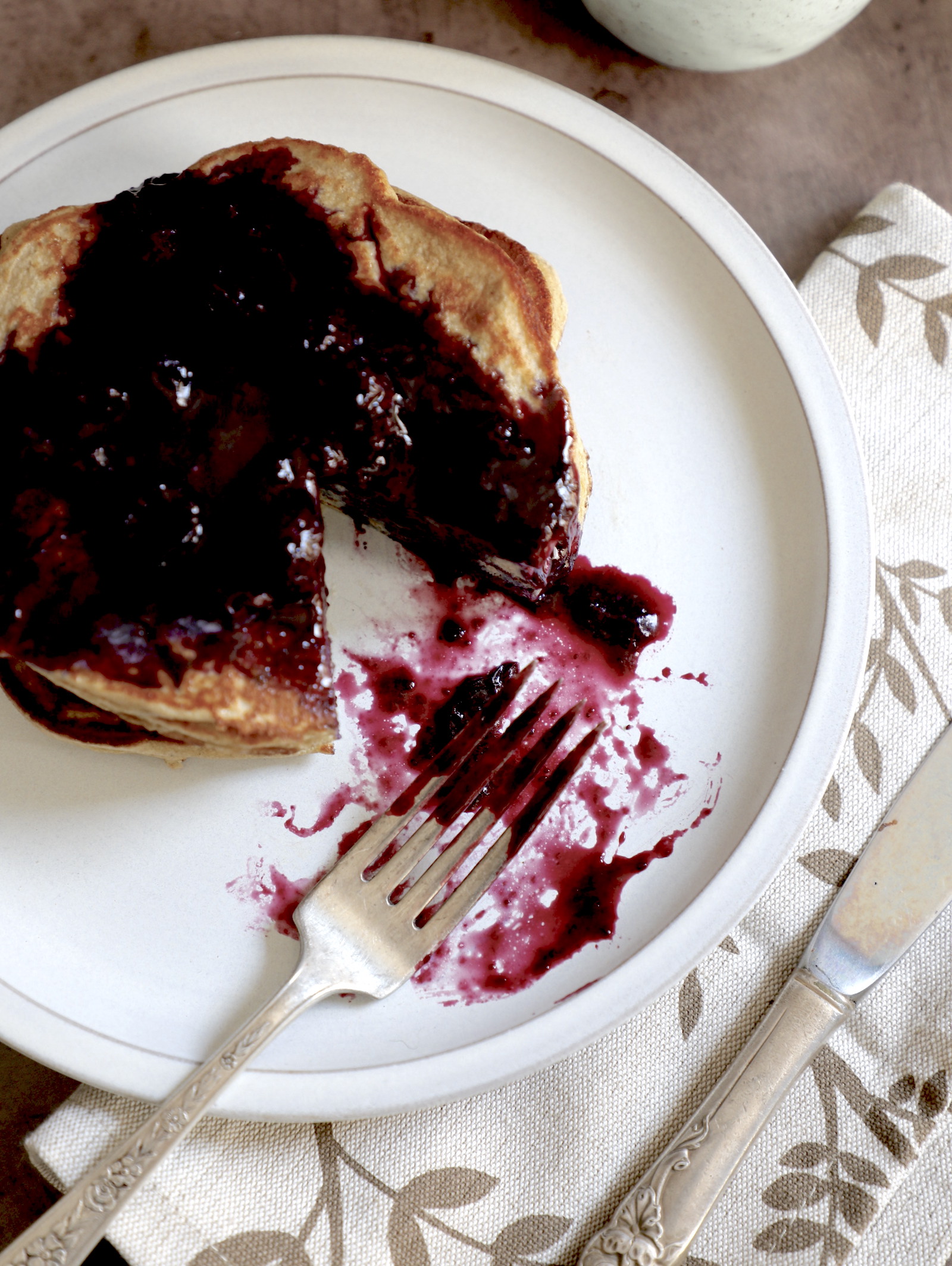 top view of pancakes on a white plate with berry compote on top and a triangle piece missing with the fork on the plate in a pool of compote.