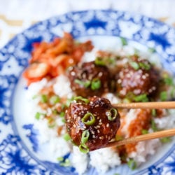 Foreground is a Korean beef bulgogi meatball held between two chopsticks with a garnish of scallions and sesame seeds. Background is a blue asian themed plate with rice and more meatballs on it.