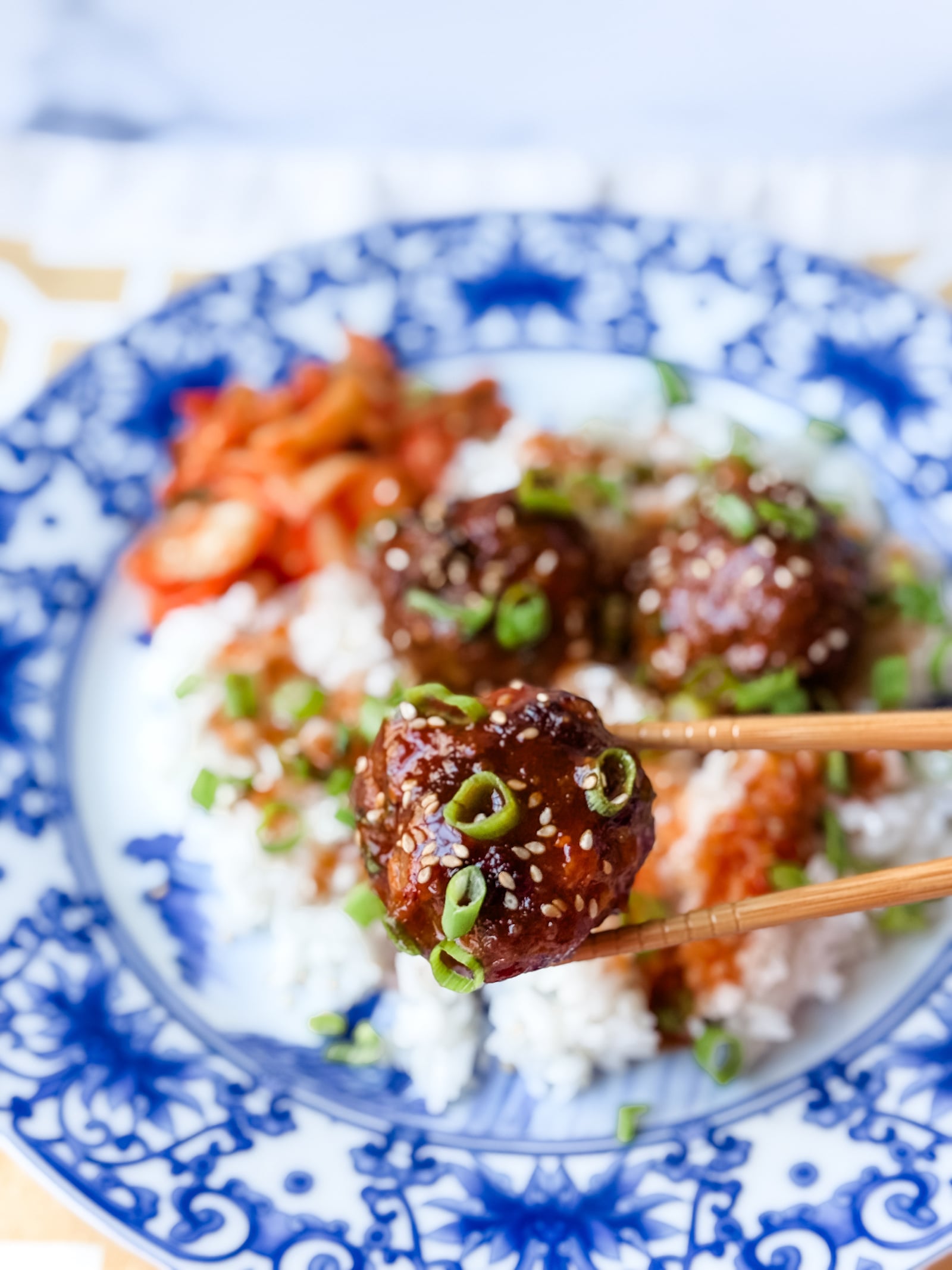 Foreground is a Korean beef bulgogi meatball held between two chopsticks with a garnish of scallions and sesame seeds. Background is a blue asian themed plate with rice and more meatballs on it.
