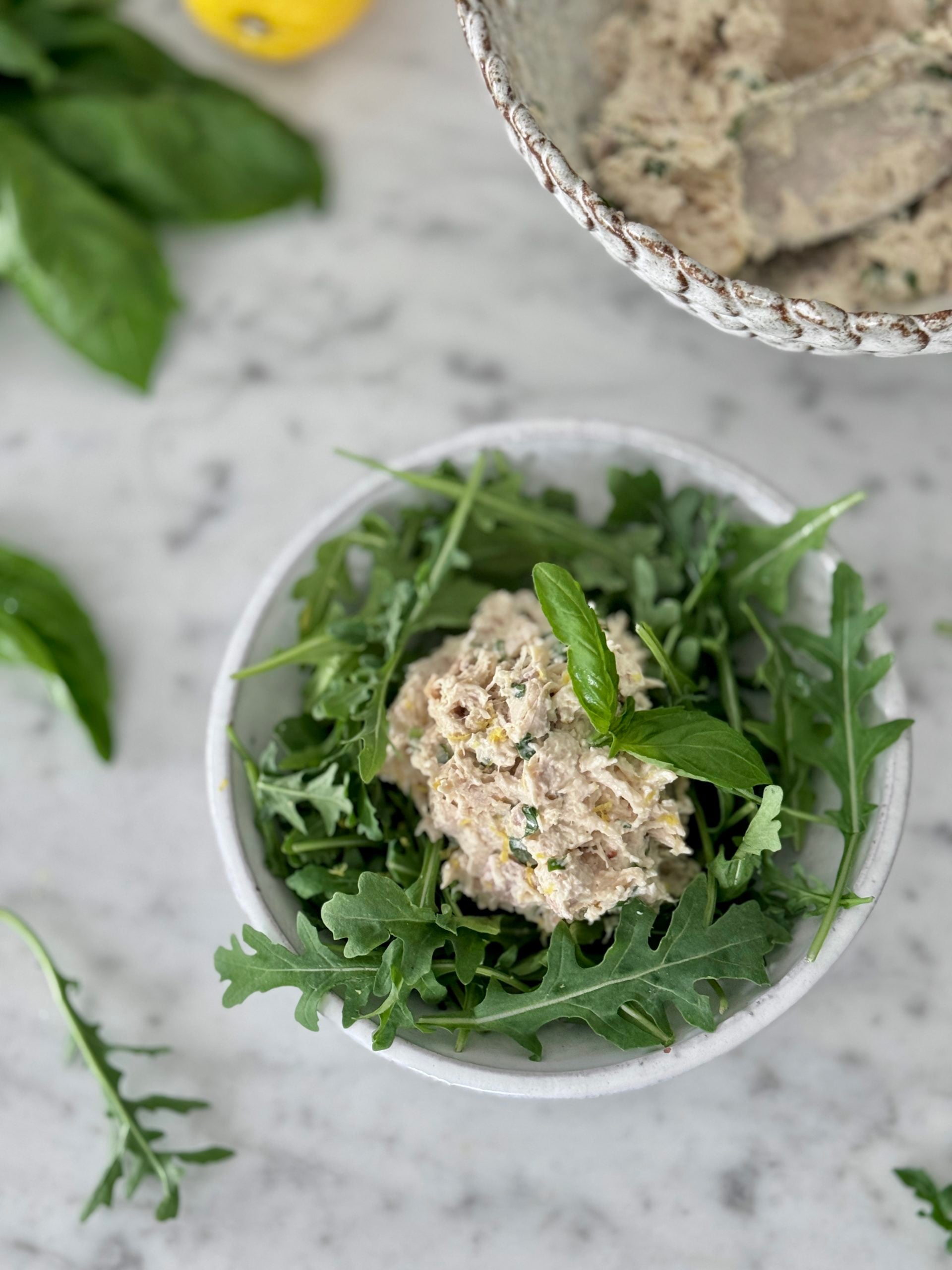 Hero shot of lemon basil chicken salad in a white bowl served on a bed of arugula and set on a marble counter with lemons, basil, and a bowl of the finished salad in the background
