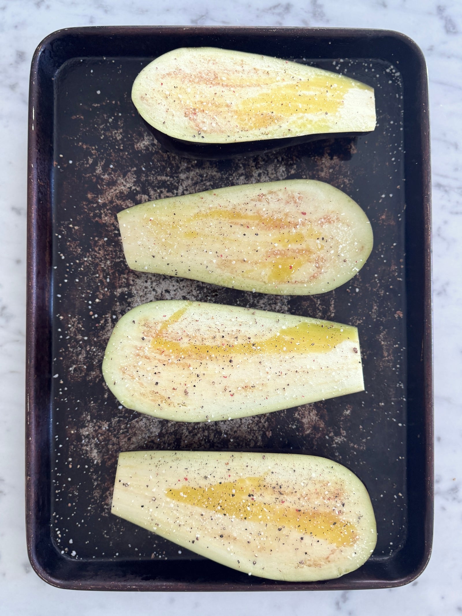 Raw eggplant halved lengthwise and drizzled with olive oil, salt and pepper, on a sheet pan ready to go into the oven for roasting